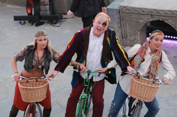 Three actors in hippie costumes riding bicycles on The Minack stage