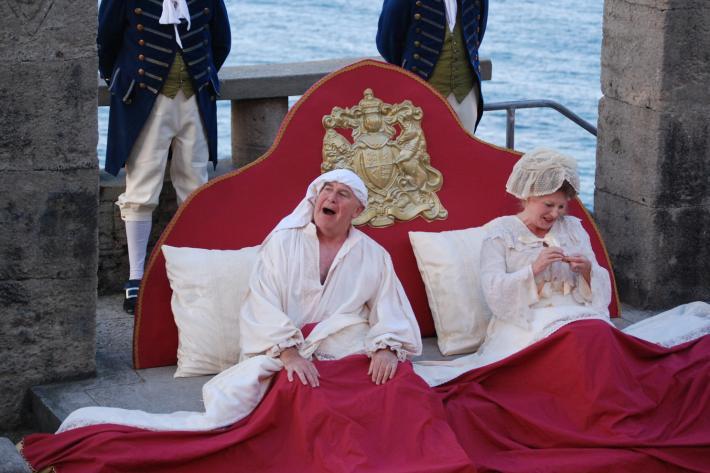 A King and Queen sit up in their royal bed on the Minack stage