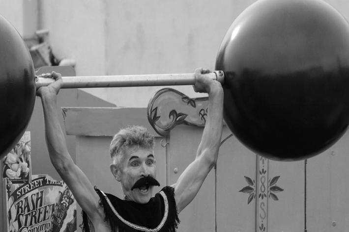 A weedy strongman lifts two enormous weights above his head.